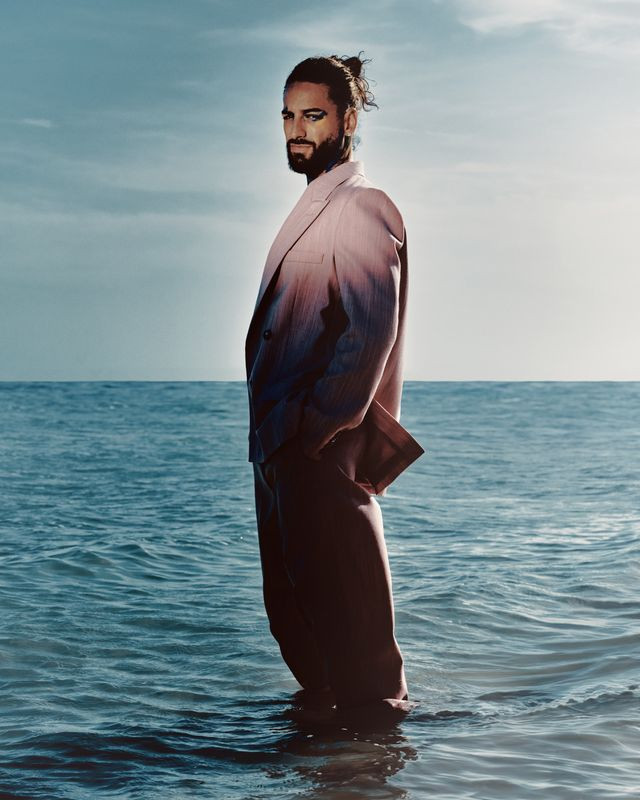 Stylish man stands in ocean with a pink ombre suit, captured in a medium shot against a backdrop of sea and sky.