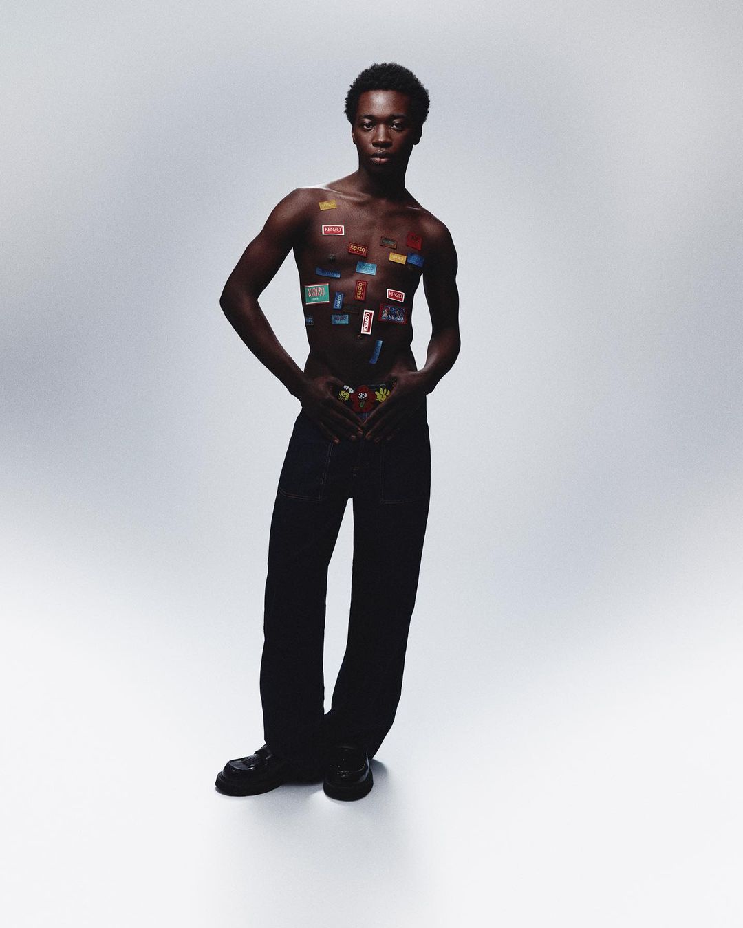 An African male fashion model poses in a studio with stickers on his chest and wearing pants and shoes against a gray backdrop.