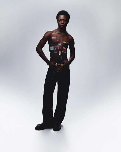 An African male fashion model poses in a studio with stickers on his chest and wearing pants and shoes against a gray backdrop.