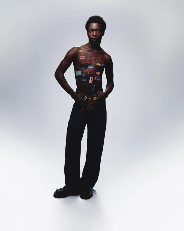 An African male fashion model poses in a studio with stickers on his chest and wearing pants and shoes against a gray backdrop.