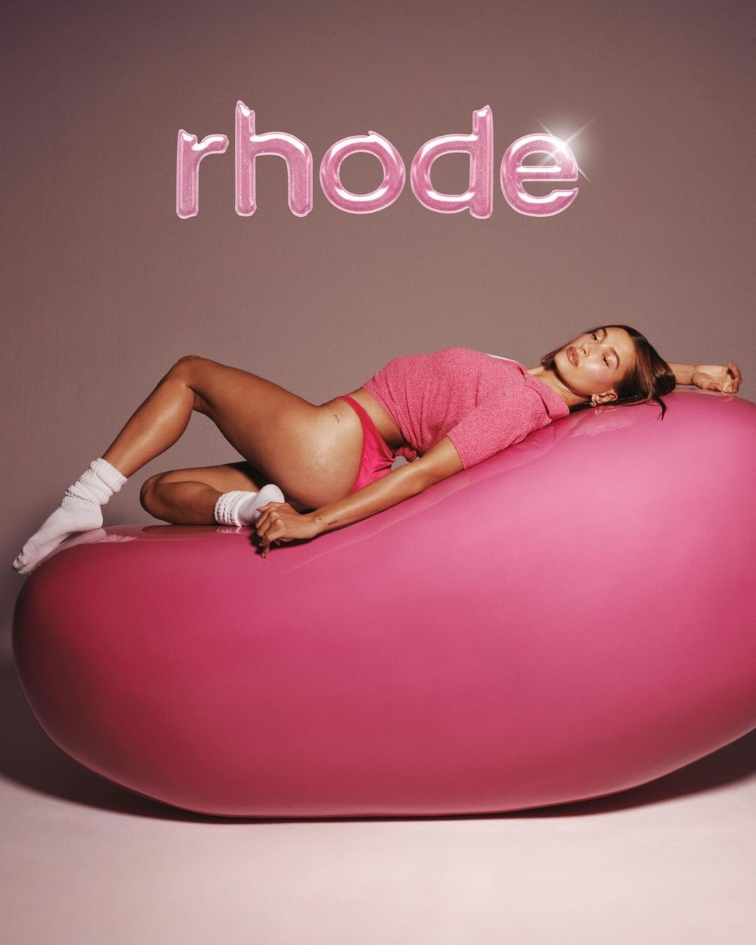 An adult female model reclines on a large pink, pill-shaped object in a studio setting, wearing a pink crop top and underwear, with the 'rhode' text logo above.