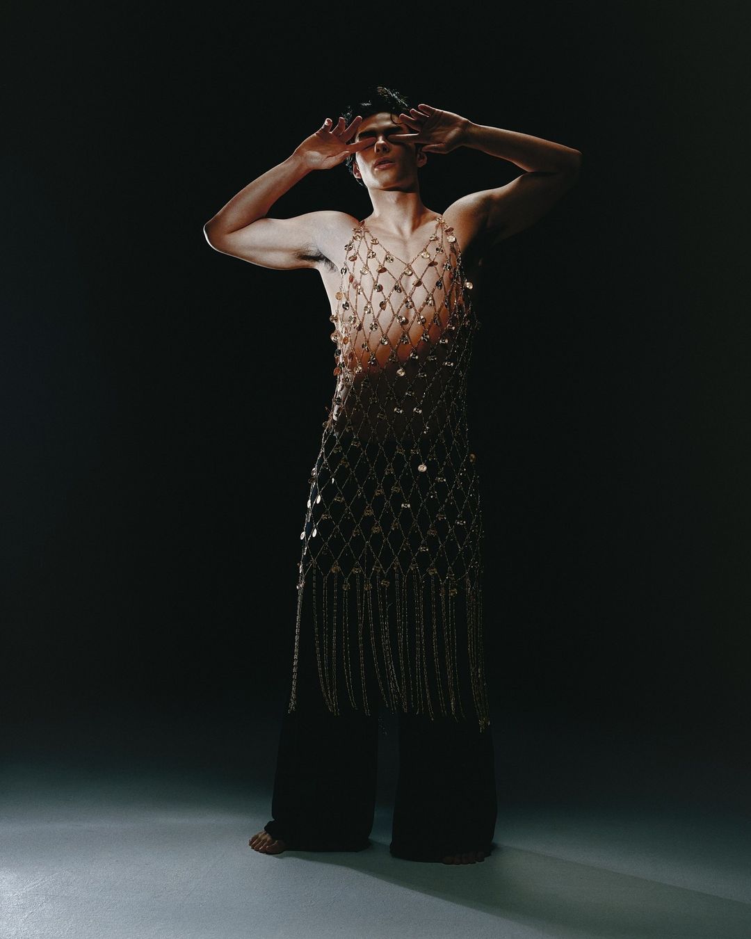 Adult male fashion model poses in net shirt and black pants, arms raised against a dark background in studio lighting.