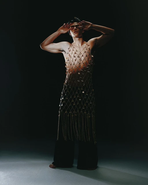 Adult male fashion model poses in net shirt and black pants, arms raised against a dark background in studio lighting.
