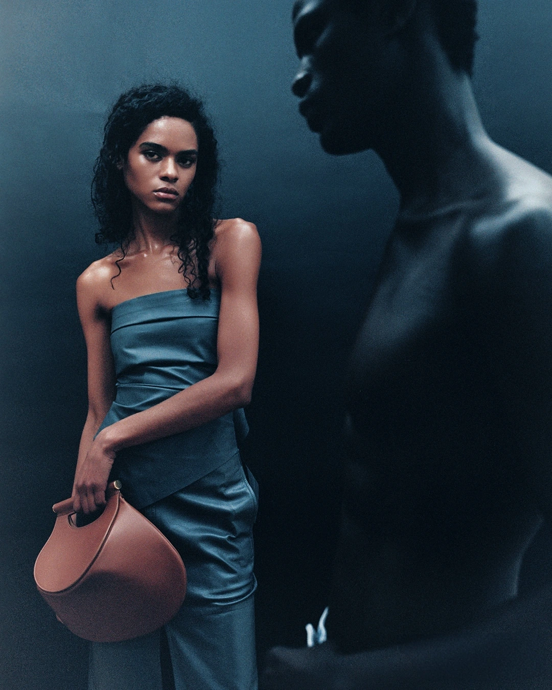 A woman in a blue dress holding a brown handbag with a man partially silhouetted in the foreground against a dark background.
