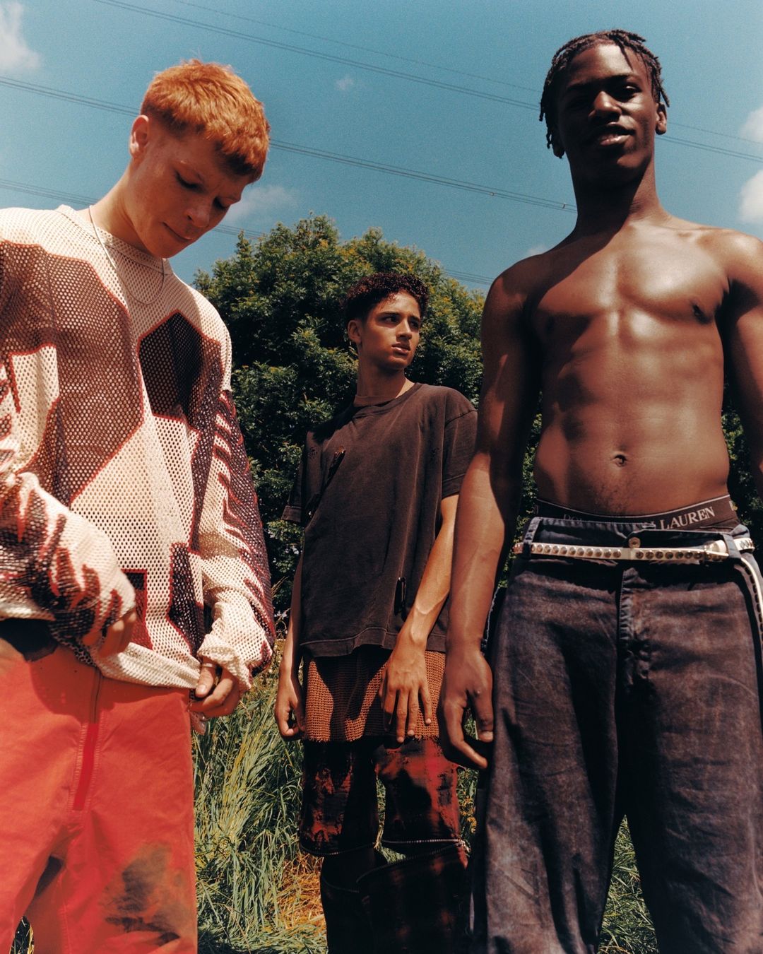 Three young men posing outdoors in low-angle sunlight against a bright blue sky and distant power lines.