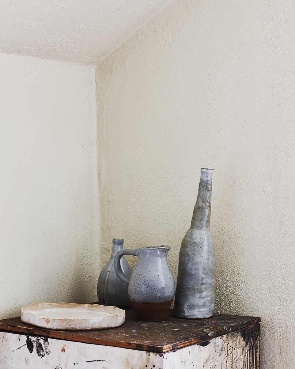 Still life composition featuring pottery vessels and plate arranged on a vintage wooden surface against a neutral wall backdrop.