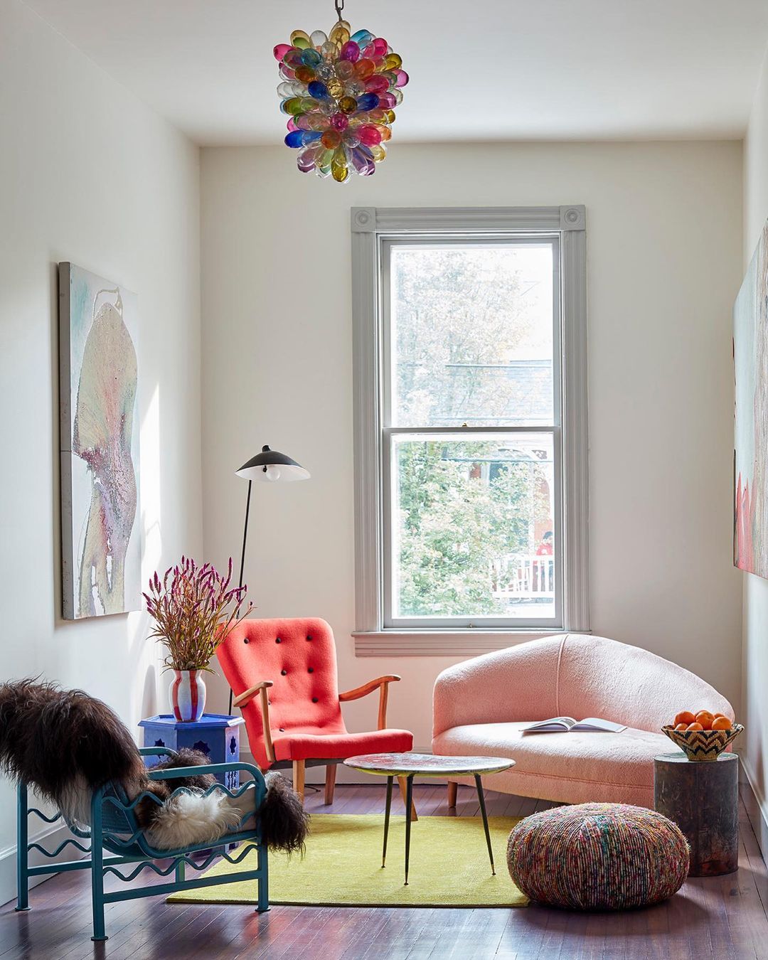 Eclectic living room interior with coral armchair, pink sofa, colorful glass chandelier, and hardwood floors in a modern home.