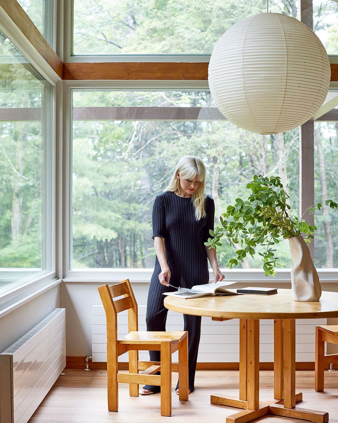 Woman reads book at round table in bright, airy room with large windows, surrounded by trees and a paper lantern pendant light.