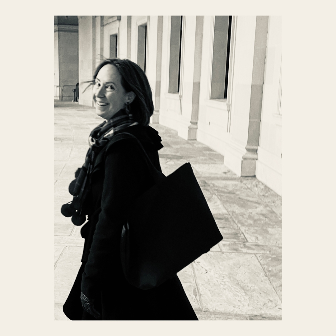 Woman in coat and scarf smiles over shoulder, carrying tote bag in colonnade setting. Black and white image with outdoor ambiance.