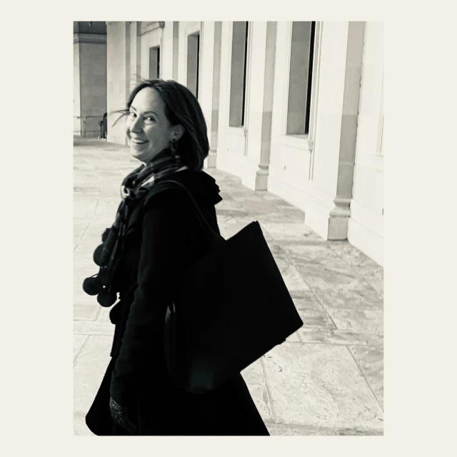 Woman in coat and scarf smiles over shoulder, carrying tote bag in colonnade setting. Black and white image with outdoor ambiance.