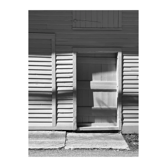 Monochrome shot of a closed plank door in a clapboard wall of a building, with a concrete walkway leading up to the doorway.