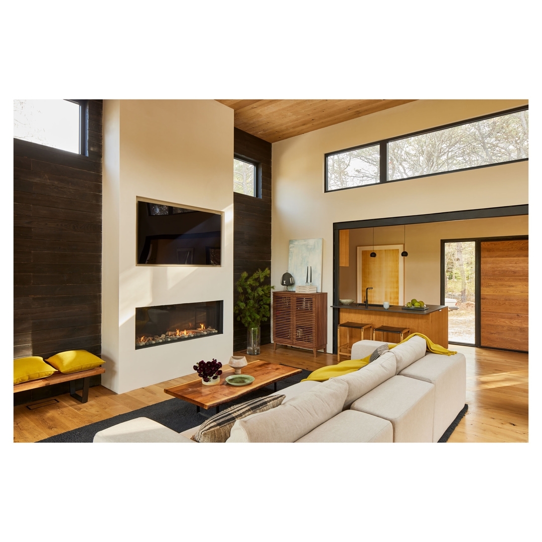 Interior of modern living room with fireplace, sofa, television, and view into kitchen; abundant natural light from large windows.