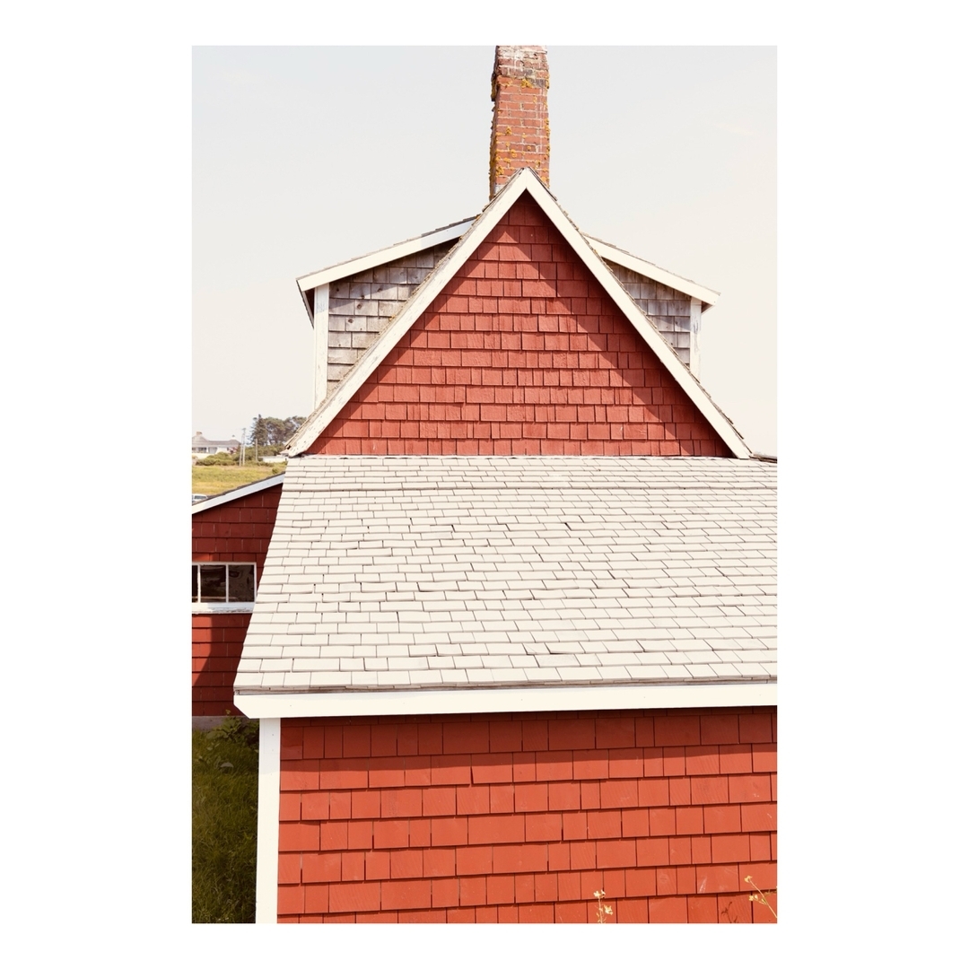 High angle view of a red clapboard house with a grey shingled roof and a brick chimney against a light blue sky and green grassy hill.