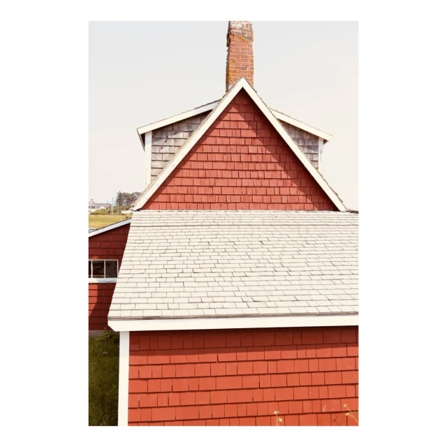 High angle view of a red clapboard house with a grey shingled roof and a brick chimney against a light blue sky and green grassy hill.