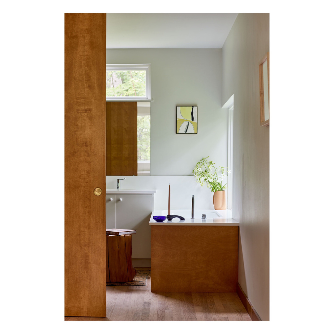 Modern bathroom interior with wood door, white cabinets, and vase of flowers, featuring a minimalist painting and natural light.