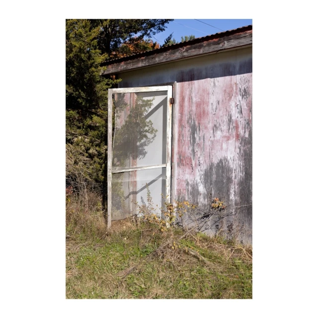 Weathered building with an open screen door surrounded by overgrown grass and trees in a rural outdoor setting on a sunny day.