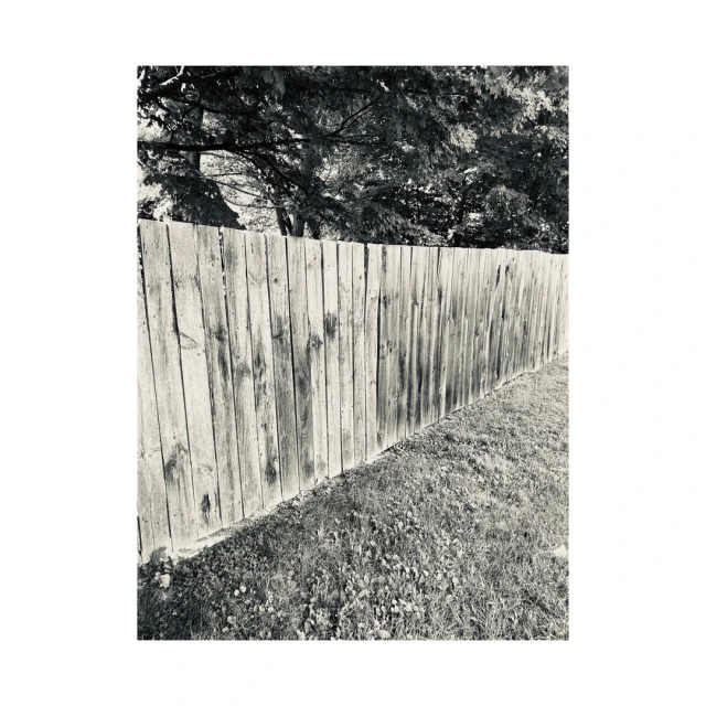 Monochrome image of a weathered wooden fence running diagonally across a grassy field, with trees visible in the background.