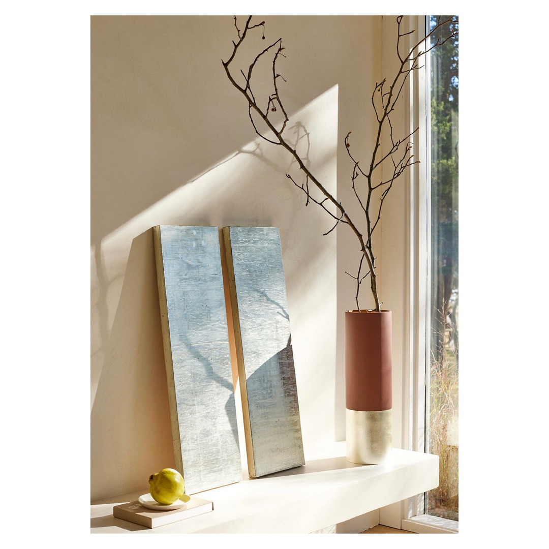 Still life featuring two silver panels, a ceramic vase with bare branches, and a yellow lemon on a small beige table indoors by a window.