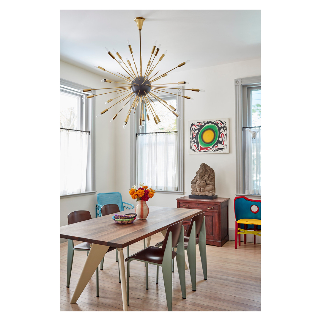 Interior of a dining room with a modern chandelier, wooden table, assorted chairs, colourful artwork, and natural light from the windows.