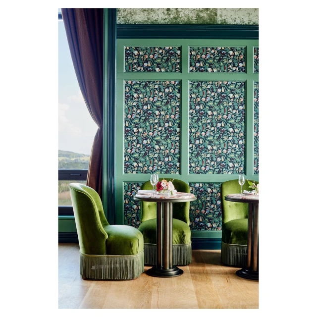 Interior of a lounge area with green velvet armchairs, round marble tables, a window, and decorative wall panels in a cozy setting.