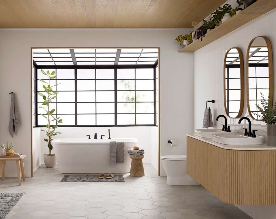 Modern bathroom interior with white bathtub, double sinks, wood vanity, geometric floor tiles, black window frame, and indoor plants.