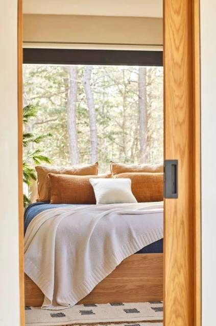 View through a doorway into a bedroom with a wooden bed frame, pillows, a cream blanket, and a forest visible through the window.