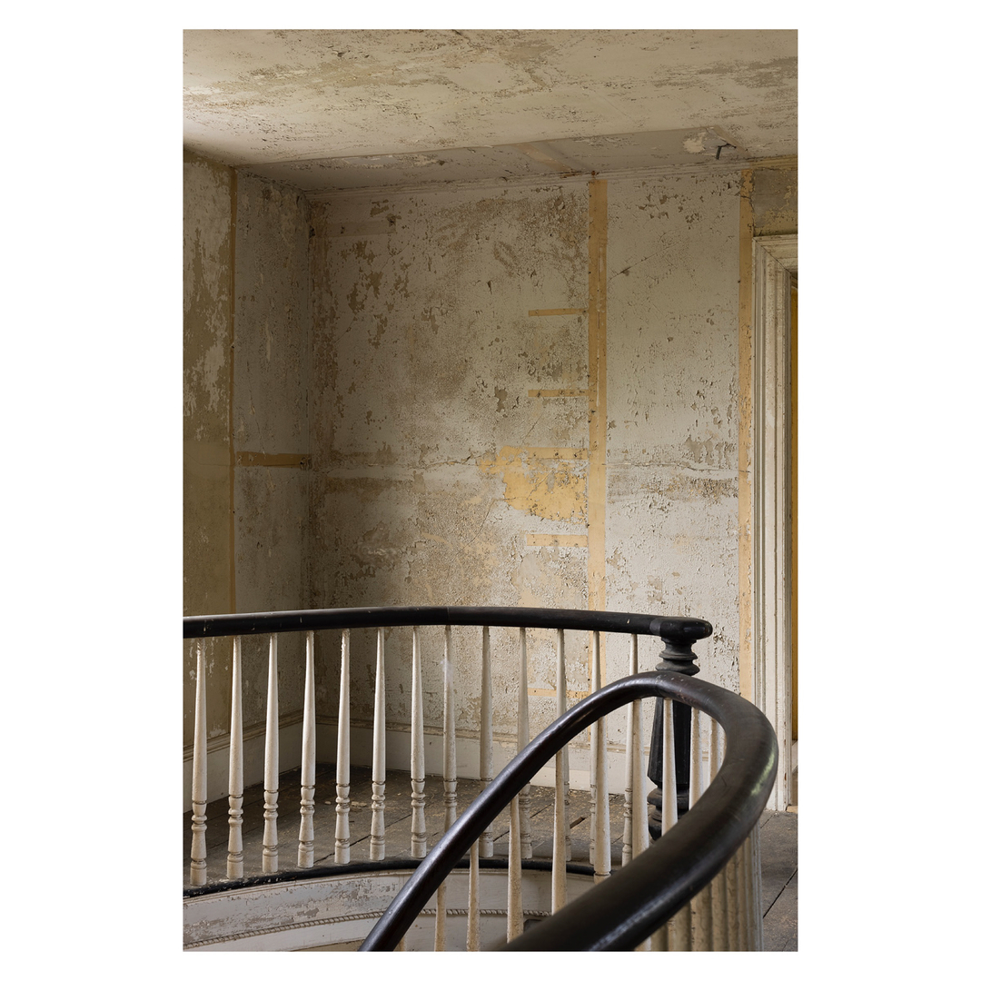 Interior view of old spiral staircase with black handrail and white spindles, peeling paint and weathered walls in a vintage home.