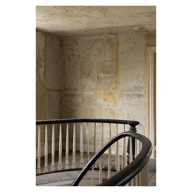 Interior view of old spiral staircase with black handrail and white spindles, peeling paint and weathered walls in a vintage home.