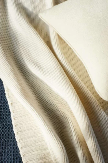 Close-up showcases a cream-colored, textured knit blanket draped alongside a matching pillow, against a deep blue knitted background.