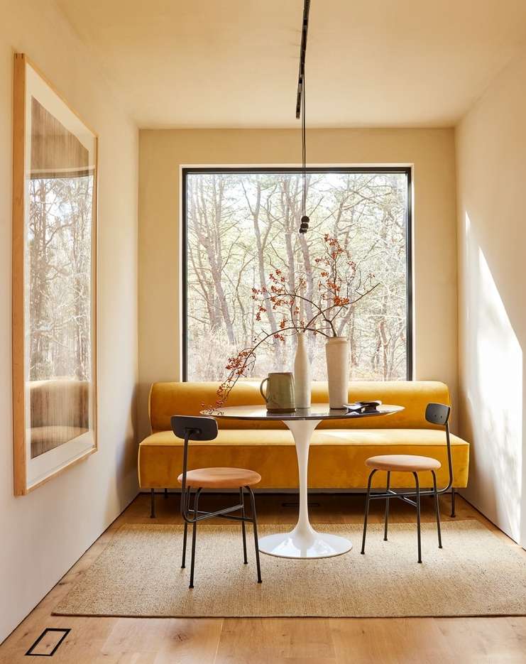 Cozy interior with a yellow sofa, round table, and two chairs by a window overlooking trees, creating a warm and inviting atmosphere.