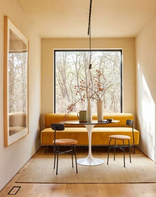 Cozy interior with a yellow sofa, round table, and two chairs by a window overlooking trees, creating a warm and inviting atmosphere.