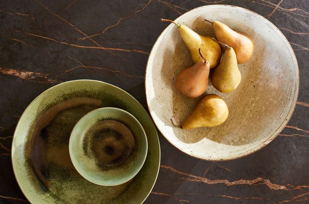 Overhead shot of pears arranged in a speckled bowl, set against a dark marble backdrop with sage green plates beside it.