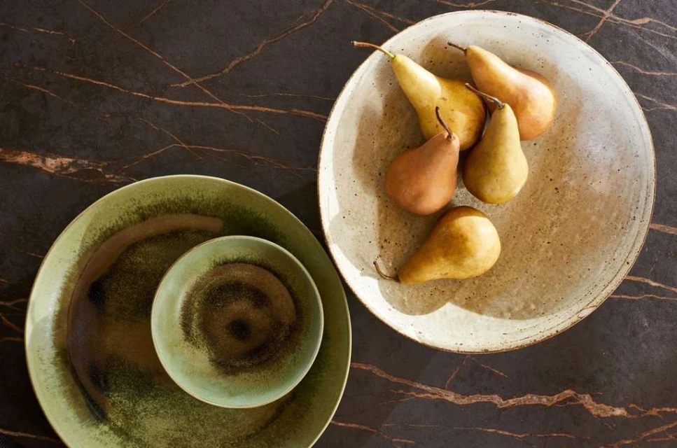 Overhead shot of pears arranged in a speckled bowl, set against a dark marble backdrop with sage green plates beside it.
