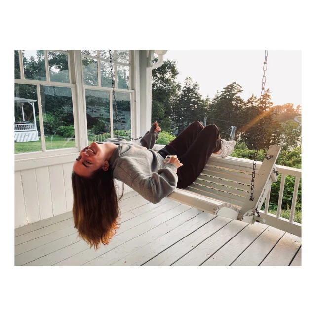 Woman on white swing on porch of white house surrounded by forest, smiling and looking up, wearing gray sweatshirt and dark sweatpants.