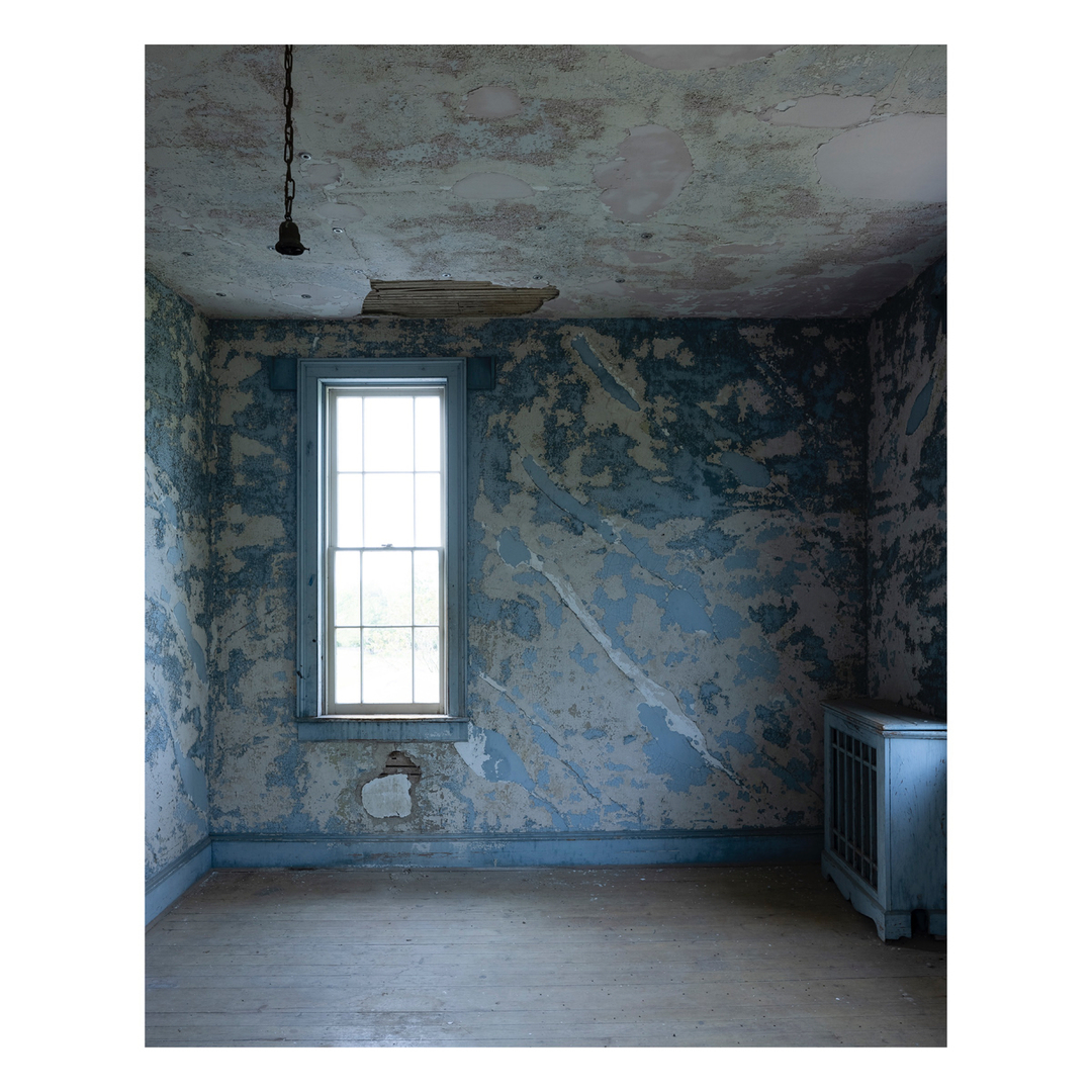 Interior of an abandoned room featuring distressed blue walls, a window with natural light, weathered wooden floor, and a hanging light fixture.