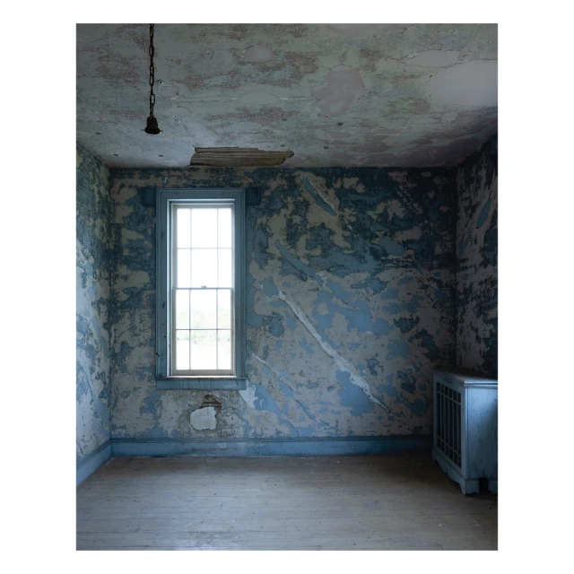 Interior of an abandoned room featuring distressed blue walls, a window with natural light, weathered wooden floor, and a hanging light fixture.