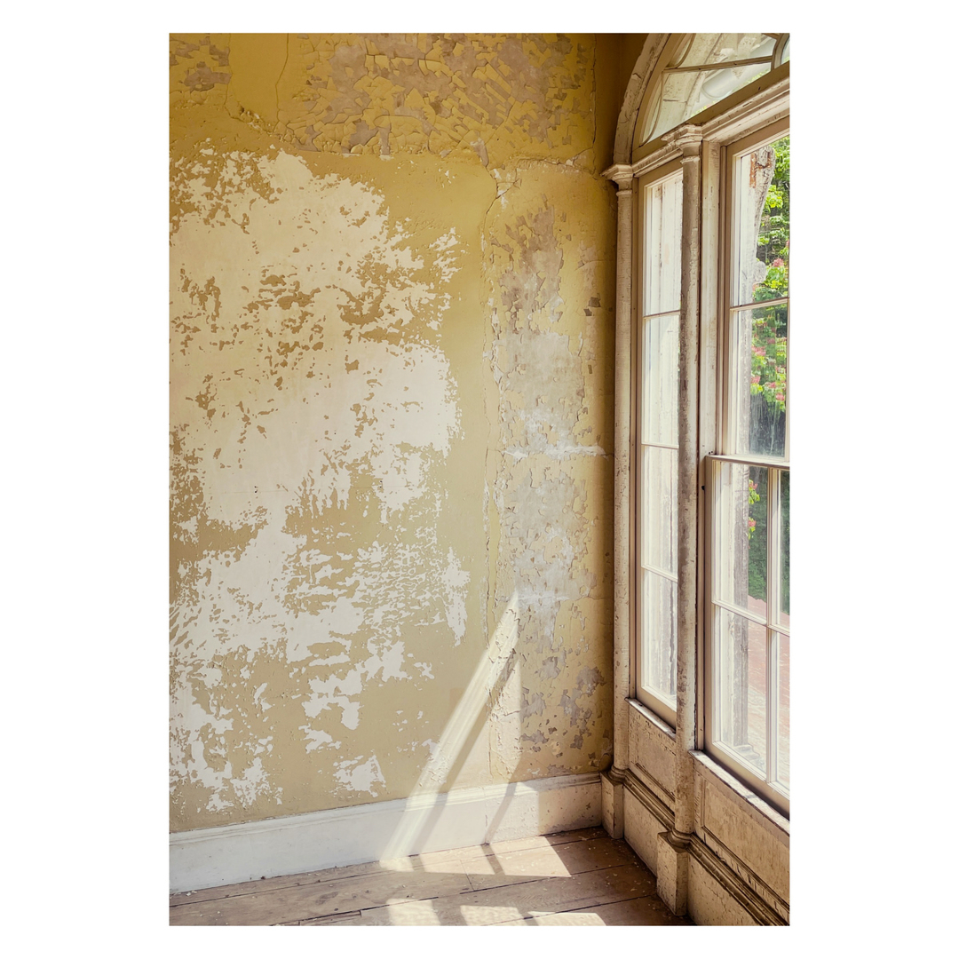 Sunlight streams through a large window illuminating a distressed yellow wall, hardwood floor, and window frame in an interior shot.