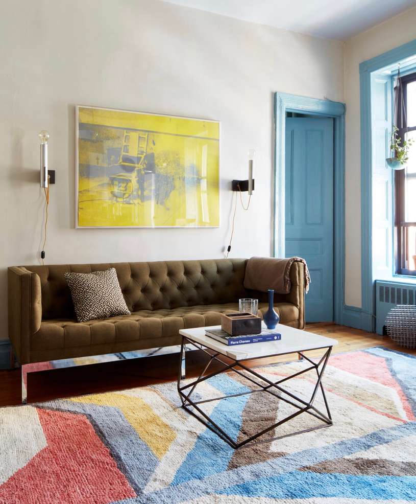 Living room interior features a tufted sofa, geometric rug, modern coffee table, and yellow abstract art against white walls.