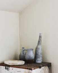 Still life photography of handcrafted ceramic vessels and a flat dish arranged on a worn wooden table against a textured white wall corner.