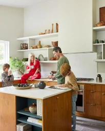 Family in a modern kitchen with wooden cabinets, a white island, and open shelving styled by photographer William Geddes.