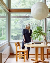 Woman in dark ribbed jumpsuit standing beside a wooden table in a bright room with large windows