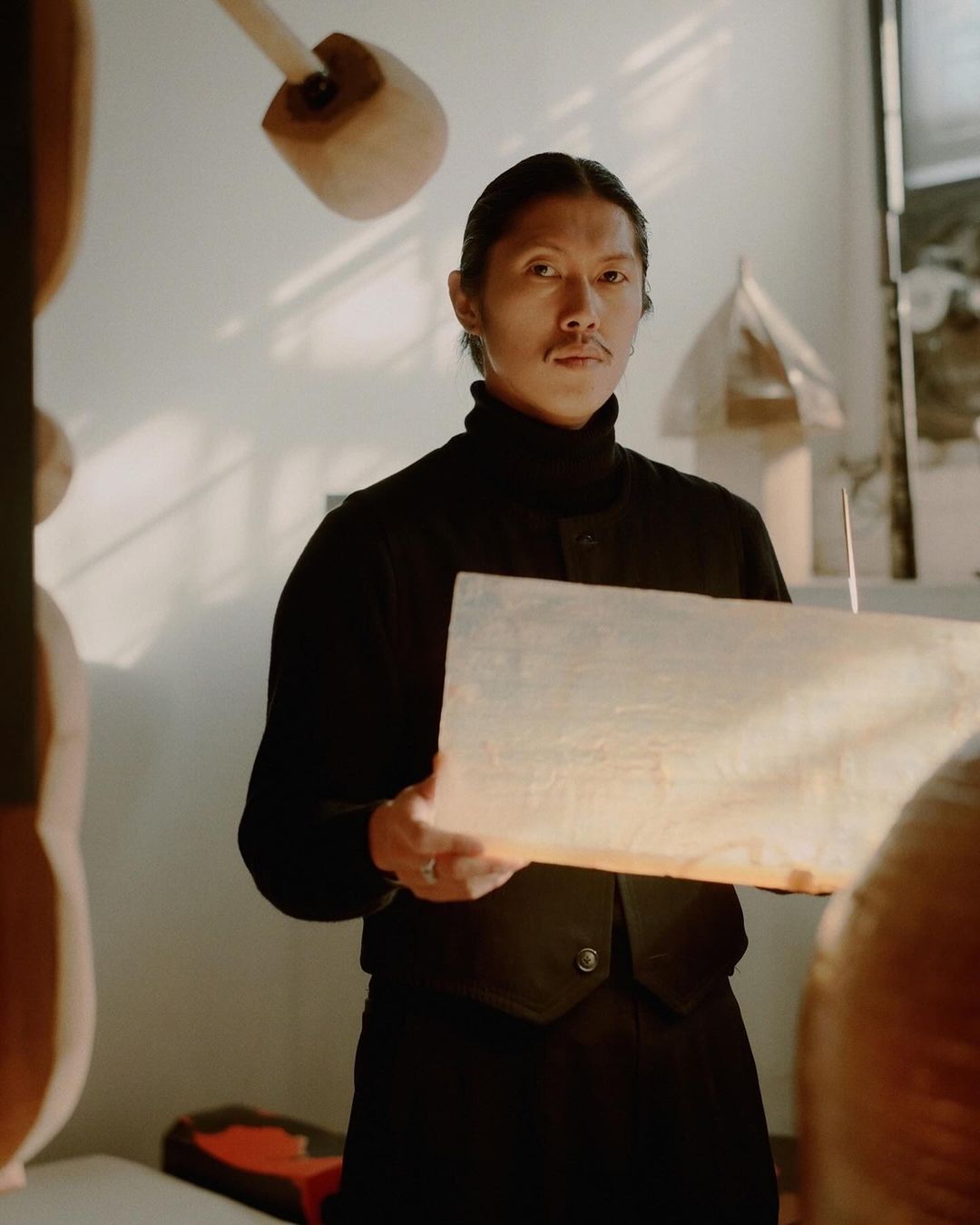 Studio portrait of a long-haired man in a black outfit, holding a glowing box. Handcrafted wooden shapes in the background.