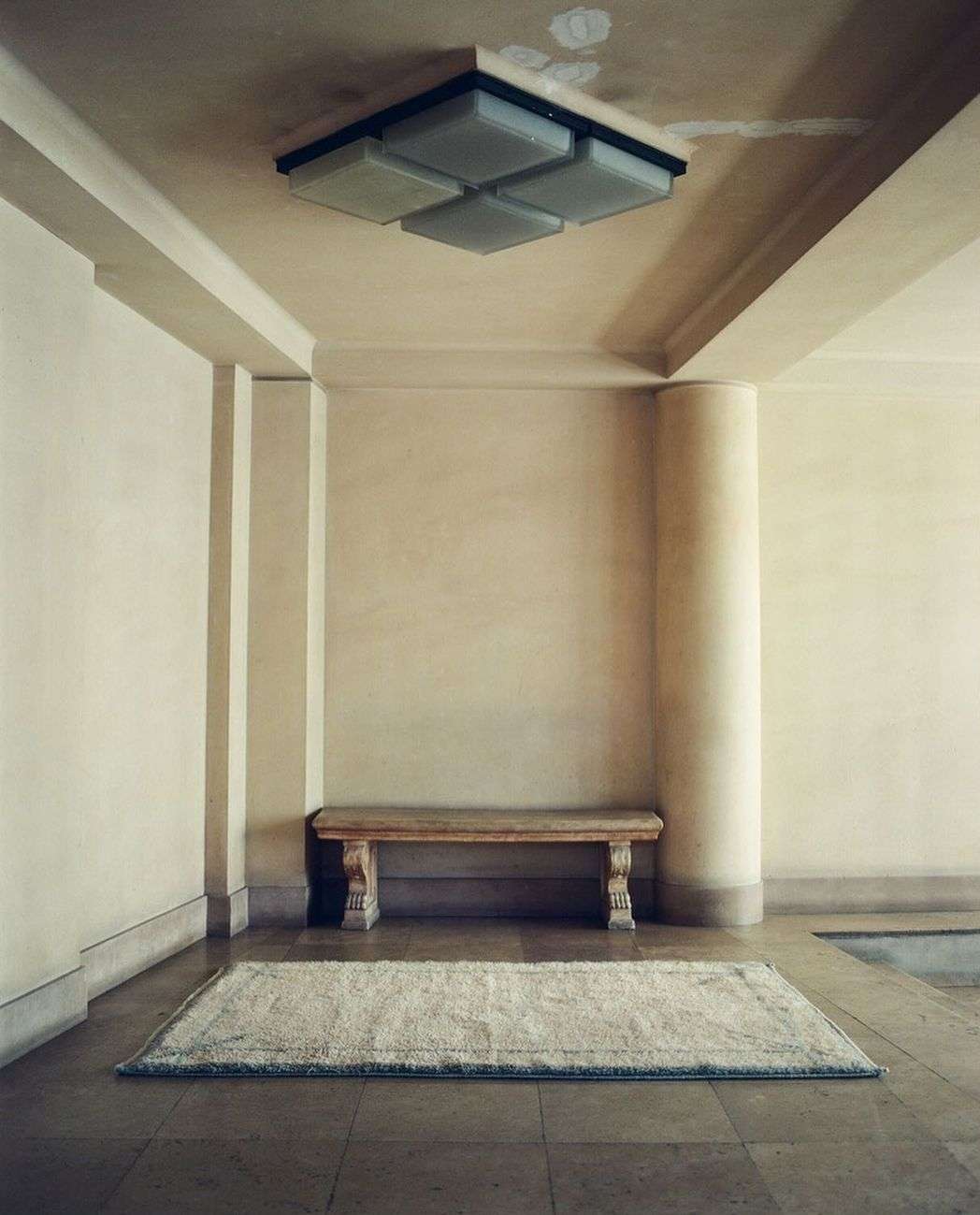 Beige interior with a stone bench and patterned rug under a modern ceiling light fixture in a spacious, neutral-toned room.
