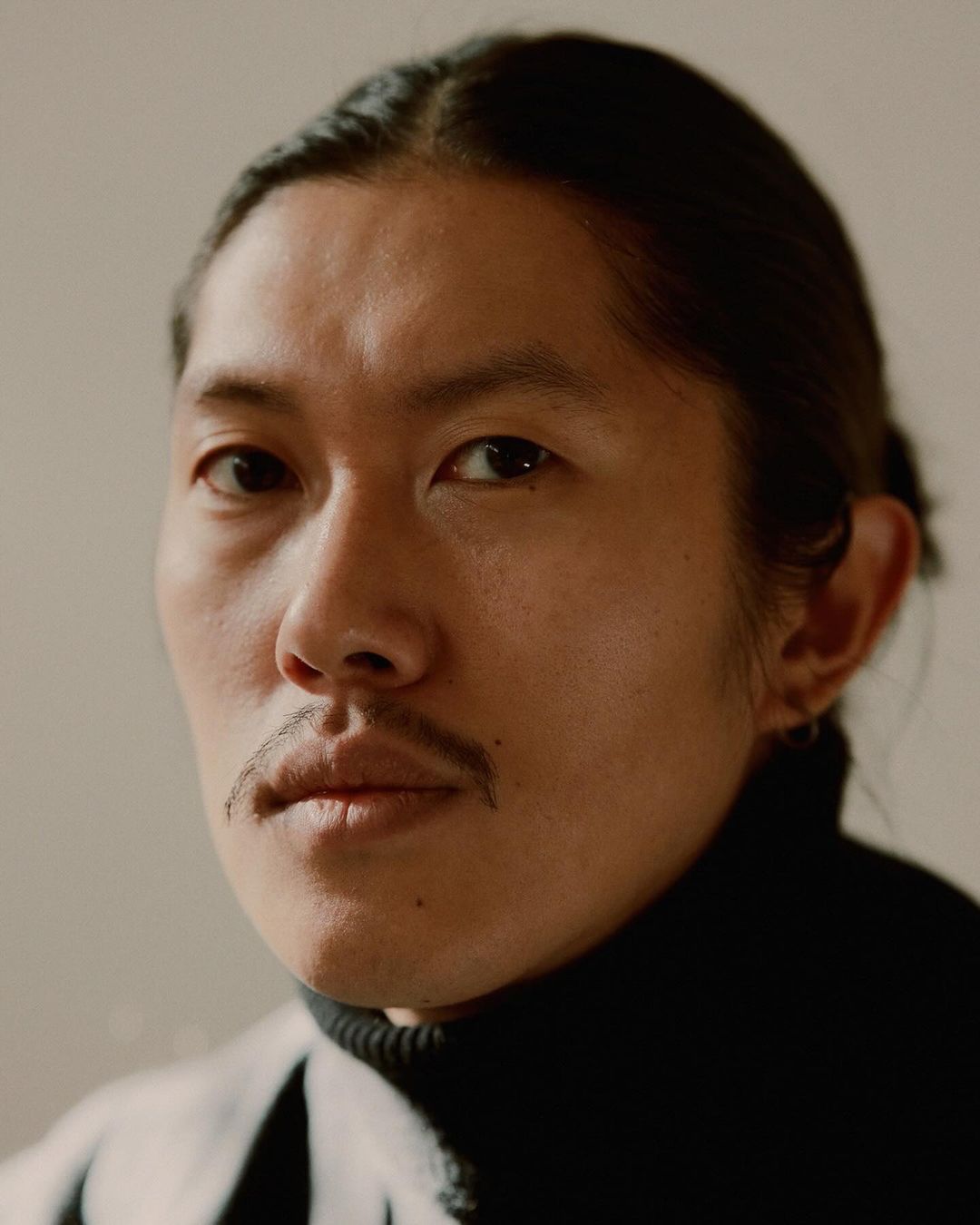 Close-up portrait of an adult male with dark hair pulled back, wearing a black turtleneck sweater against a neutral-toned background.
