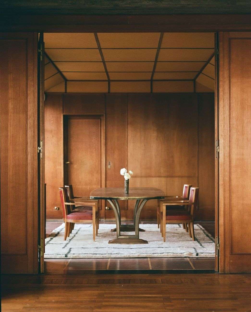 Interior view of a dining room featuring a wooden table set with white flowers, surrounded by chairs on a striped rug.