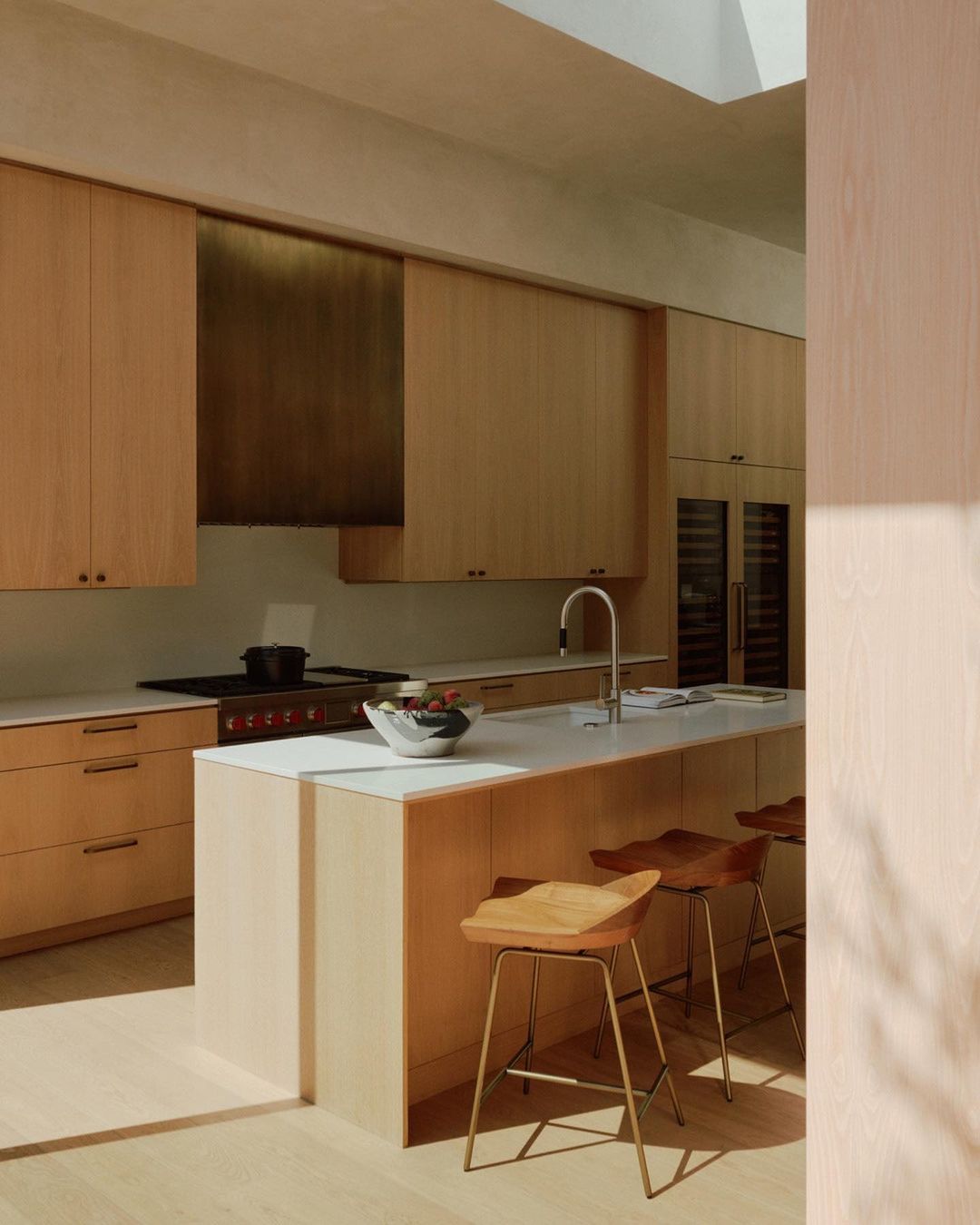 Light wood kitchen with island, brass accents, and three wooden stools in a minimalist, modern design, illuminated by soft natural light.