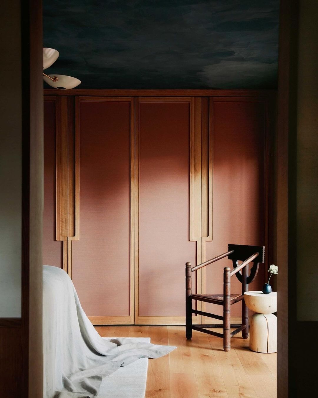 Interior view of a bedroom with a wooden chair, side table and bed, featuring wood flooring, paneled walls, and a dark blue ceiling.