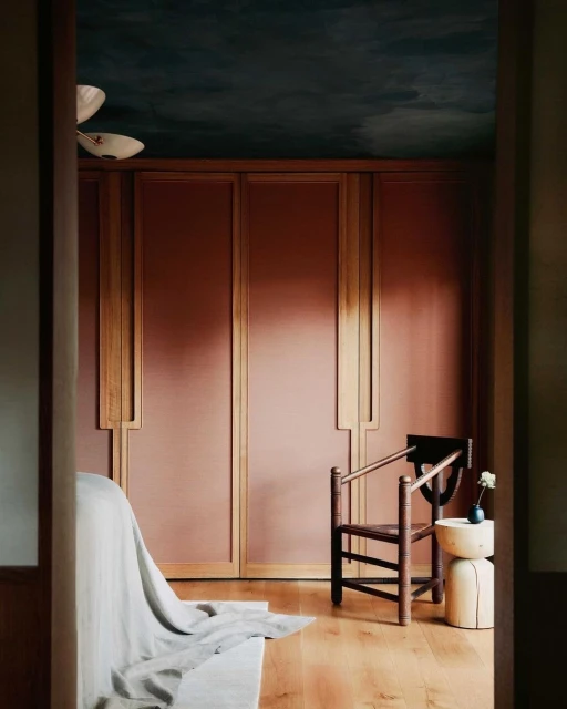 Interior view of a bedroom with a wooden chair, side table and bed, featuring wood flooring, paneled walls, and a dark blue ceiling.