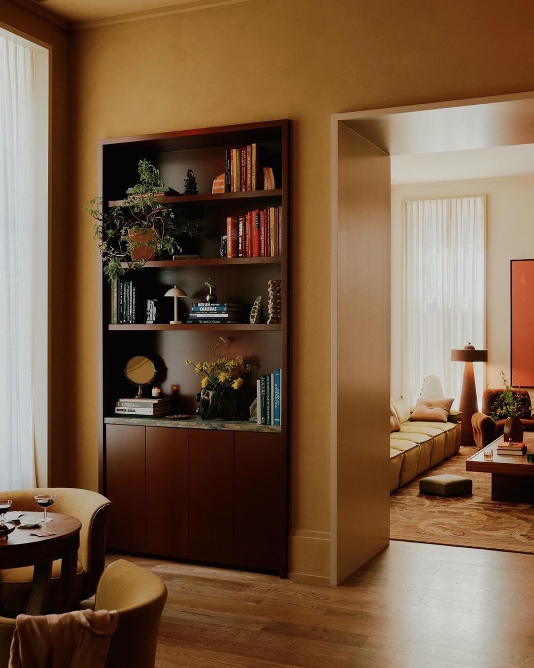 Well-lit interior of a living room features a bookshelf filled with books and decorative items, complemented by seating and warm color tones.