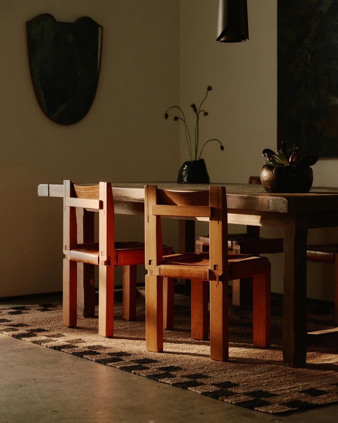 Warm interior shot of a rustic wooden dining table with chairs, vase with flower stems, dark lamp shade, and shield wall decor.